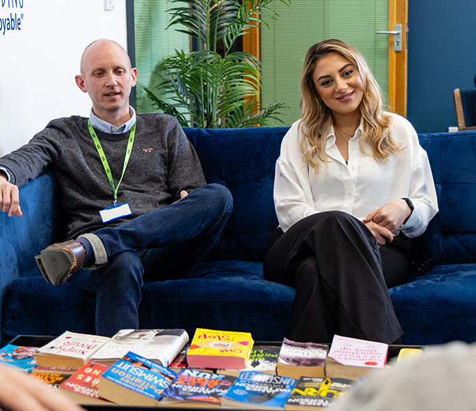 A man and a woman sit on a blue sofa in an office, with various books laid out on a coffee table in front of them.
