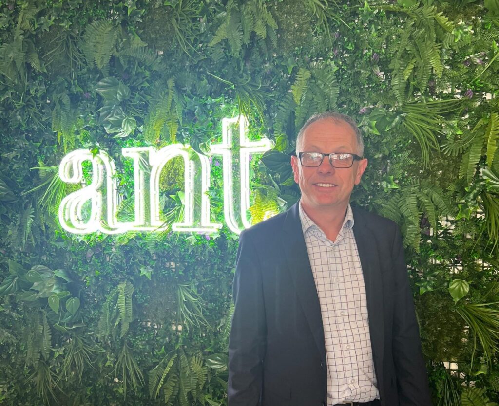 Chris Forrest, wearing a suit and glasses, stands in front of a green wall adorned with plants and a neon sign that reads 'ant'.